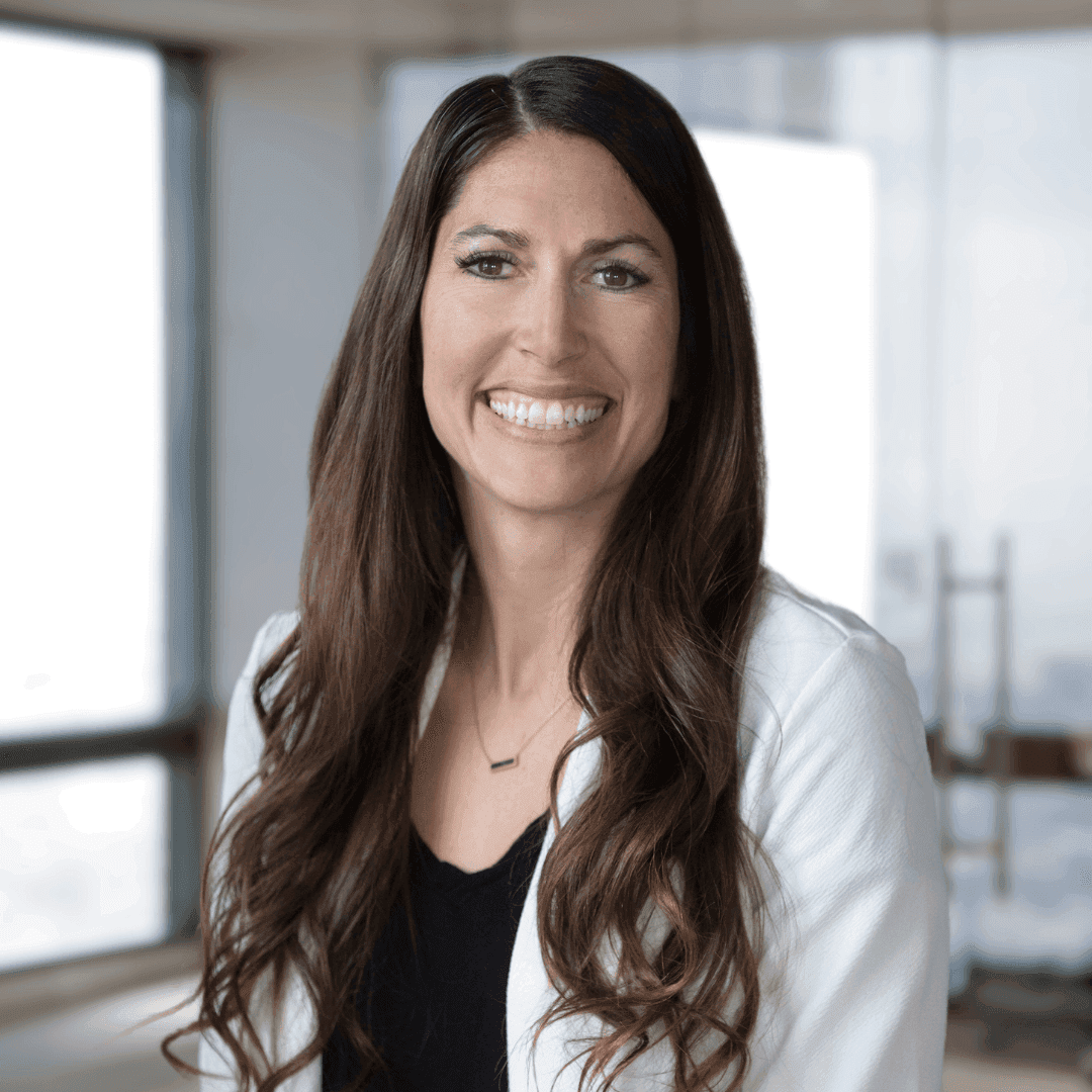 Person with long brown hair wearing a white blazer and black top, standing in an office space.