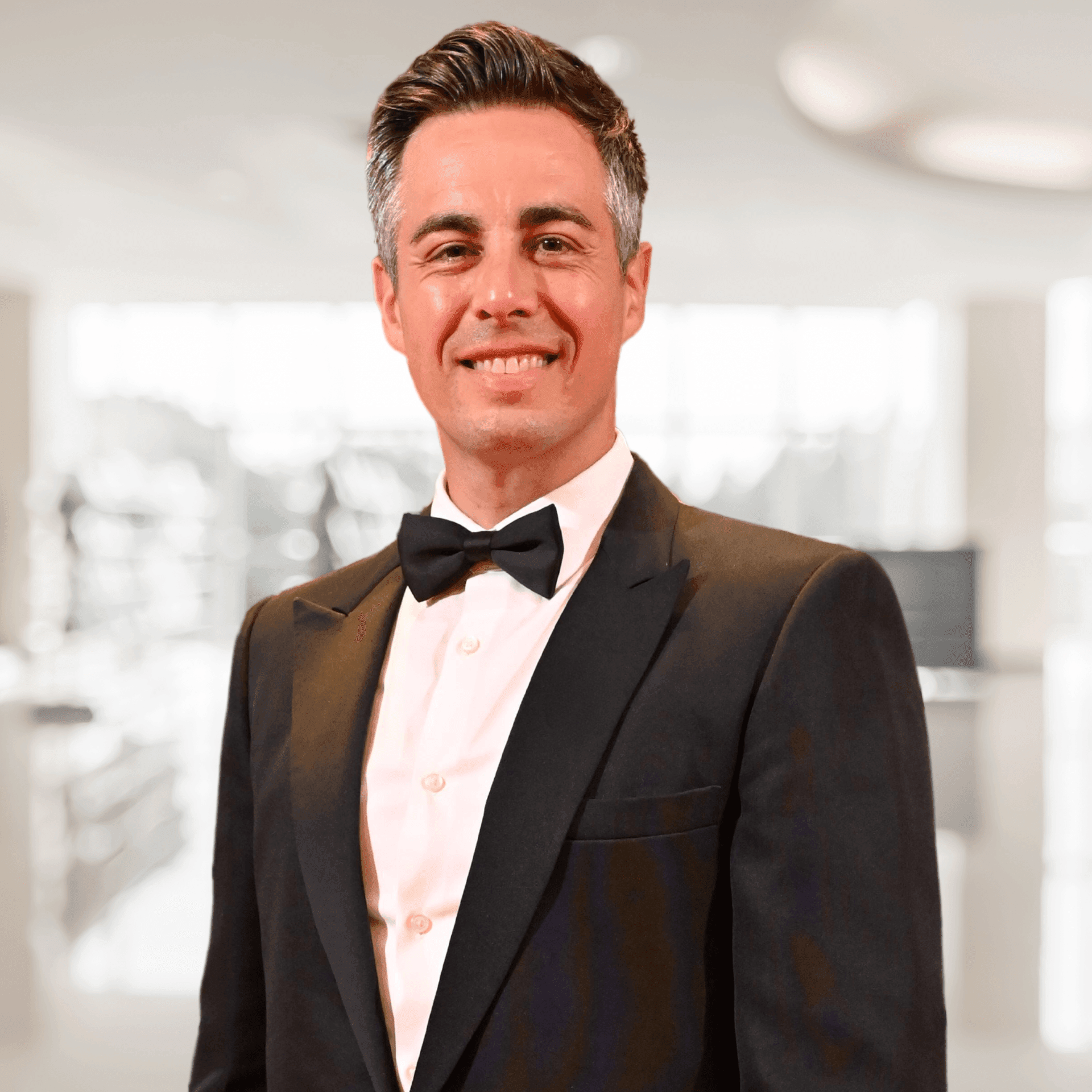 Man in a tuxedo smiling in a bright indoor setting with blurred background.