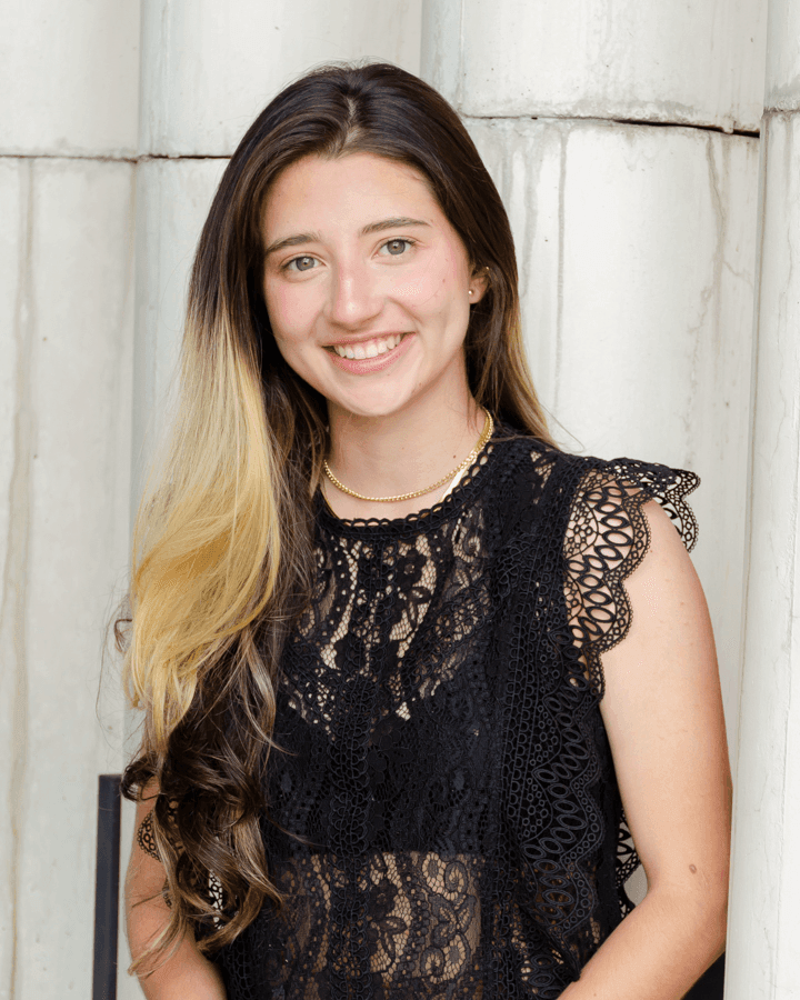 Woman wearing a black lace top with gold jewelry, standing in front of white pillars.